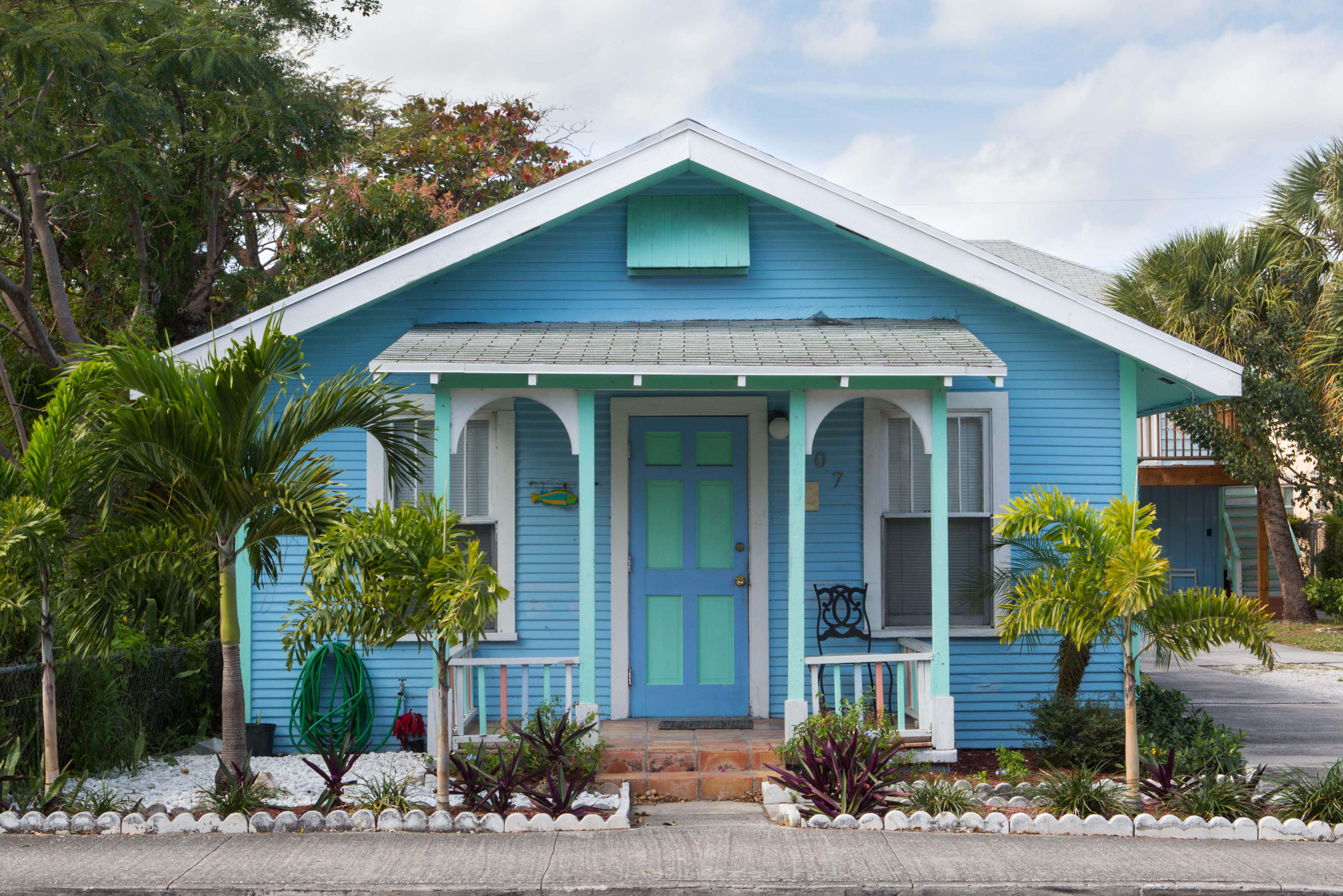 A charming blue house with white trim features a turquoise front door and decorative landscaping. Palm trees and colorful plants surround the entrance, under a partly cloudy sky.