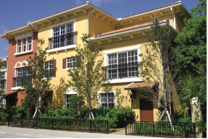 A vibrant two-story townhouse with a mix of yellow and reddish-brown exterior. It features multiple windows with decorative black railings, surrounded by small trees and shrubs, and has a decorative fence at the front. The sky is clear and blue.