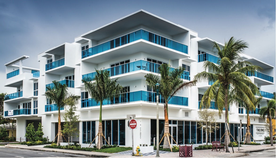 A modern, multi-story building with white walls and large blue glass balconies. Several palm trees are in front, adding to the tropical atmosphere. The scene features a clear sky and a stop sign nearby.
