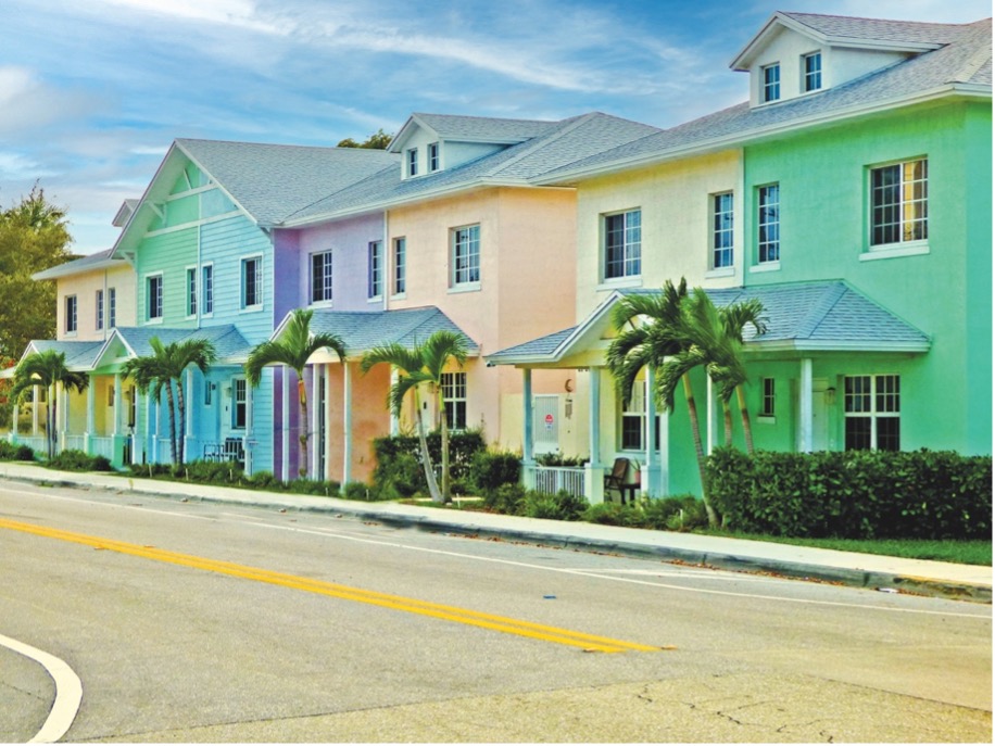 Colorful buildings line a street, featuring pastel hues of pink, green, and blue. Lush palm trees are planted along the sidewalk, complementing the vibrant architecture. A clear sky adds to the cheerful atmosphere.