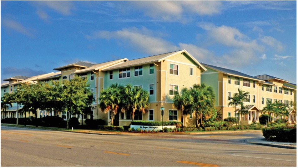 A multi-story residential building with a light green exterior, surrounded by palm trees and landscaping, under a blue sky with some clouds. A sidewalk and road run in front of the building.
