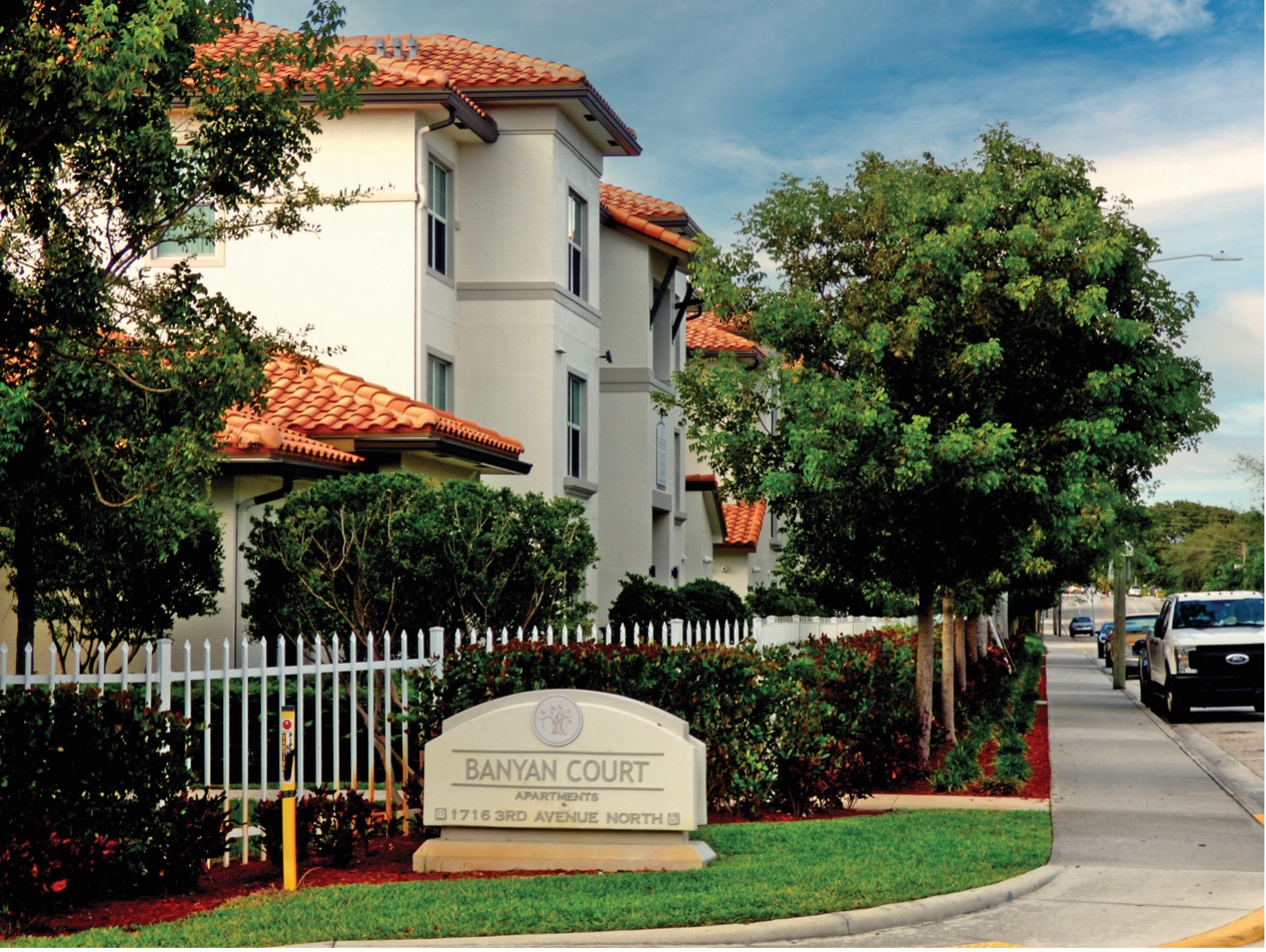 A residential apartment complex named Banyan Court, featuring two-story buildings with red tile roofs and manicured landscaping. A white fence surrounds the property, and a sign displays the complex's name. A street with parked cars runs alongside the complex.