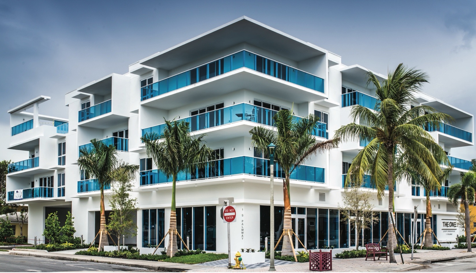 Modern multi-story building featuring white exterior and blue glass balconies, surrounded by palm trees. The structure has large windows and a contemporary design, set against a cloudy sky.