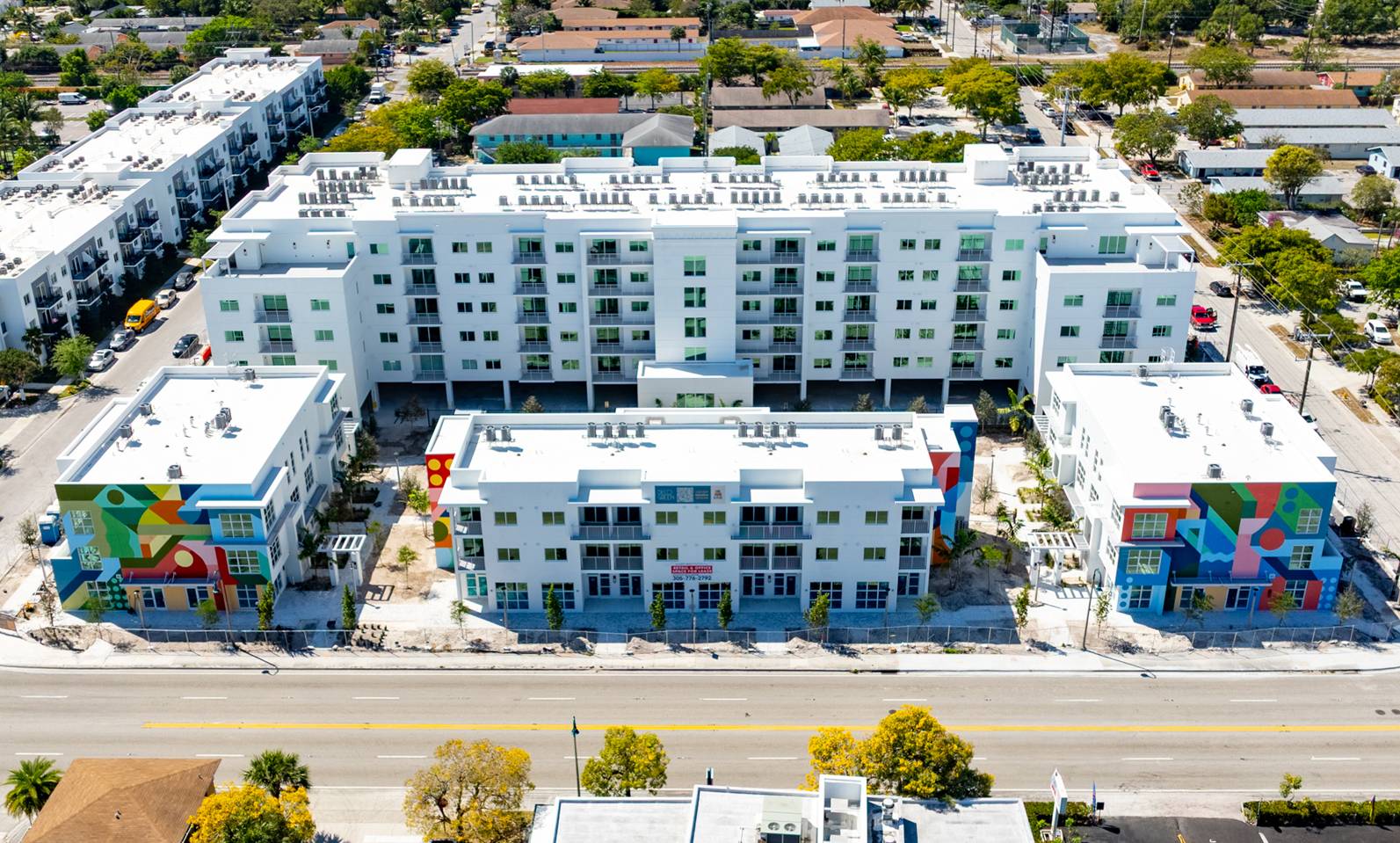 Aerial view of a modern, multi-story residential complex featuring colorful murals on the lower floors, surrounded by lush trees and landscaping. The building has a flat roof and multiple balconies, with a street visible in front.