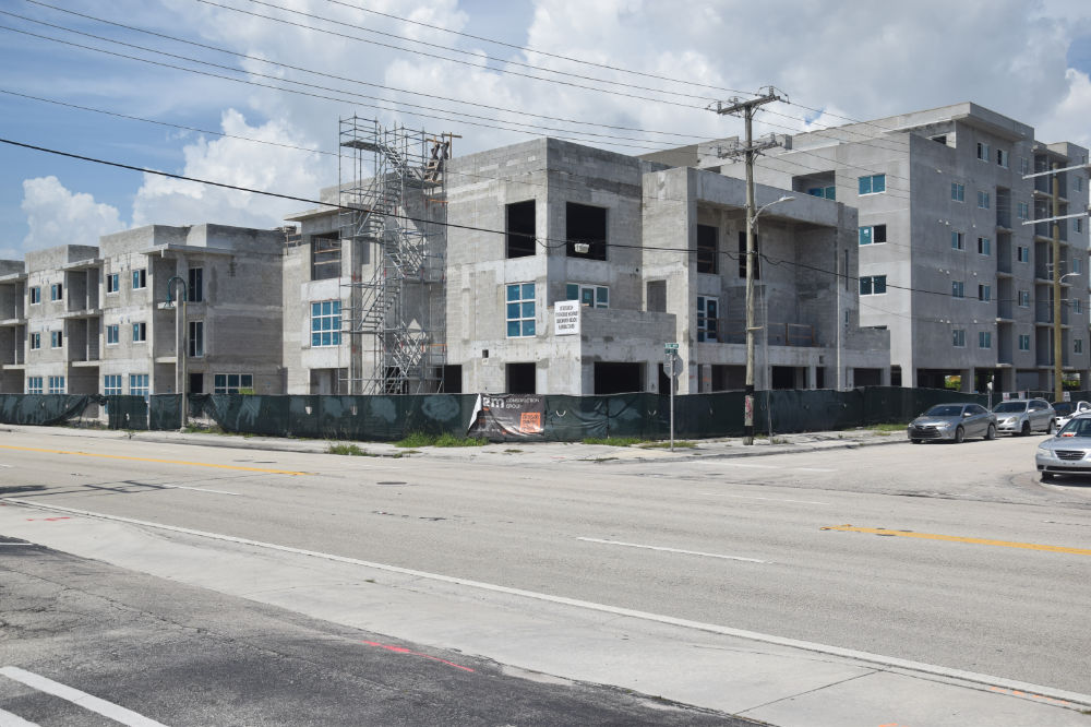 Newly constructed multi-story buildings with unfinished exteriors, scaffolding, and large windows under a partly cloudy sky, located along a road with few vehicles.