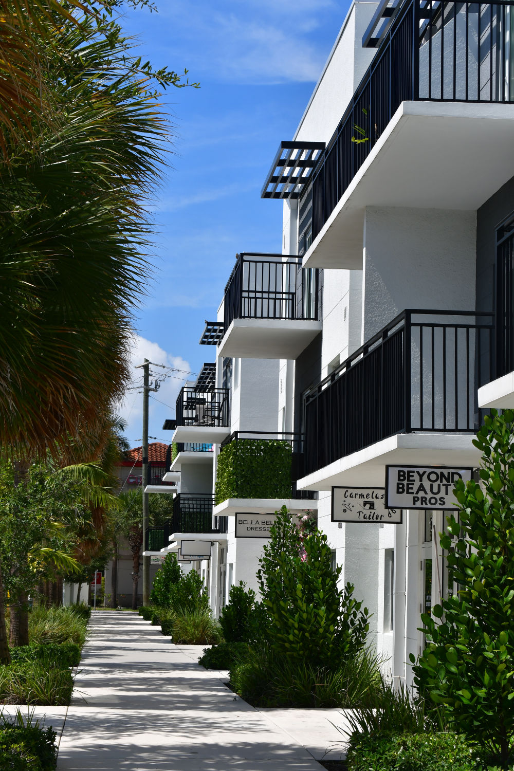 Modern multi-story buildings line a walkway, featuring balconies and lush greenery. The sky is bright with a few clouds, enhancing the vibrant atmosphere.